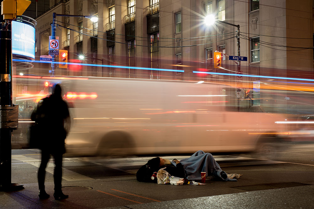 Homeless man on the streets of Toronto on a cold night with moving people and cars showing poverty in the city.