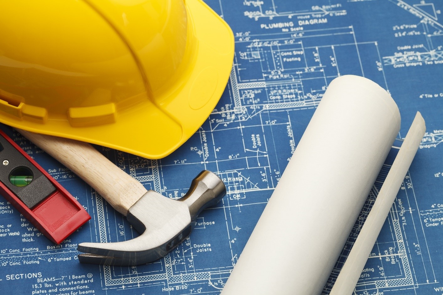 A yellow hard hat sits on top of blueprints for a building. Also seen in the photo beside the hat is a hammer and a level, and a rolled up sheet of more blueprints.